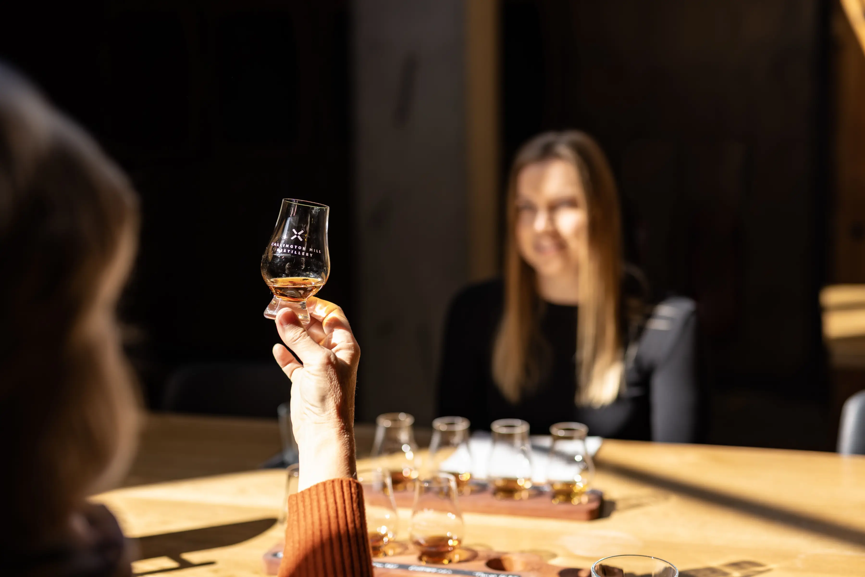 A hand holding a Callington Mill Distillery whisky tasting glass filled with amber spirit, with a tasting flight of several glasses on a wooden board blurred in the background.