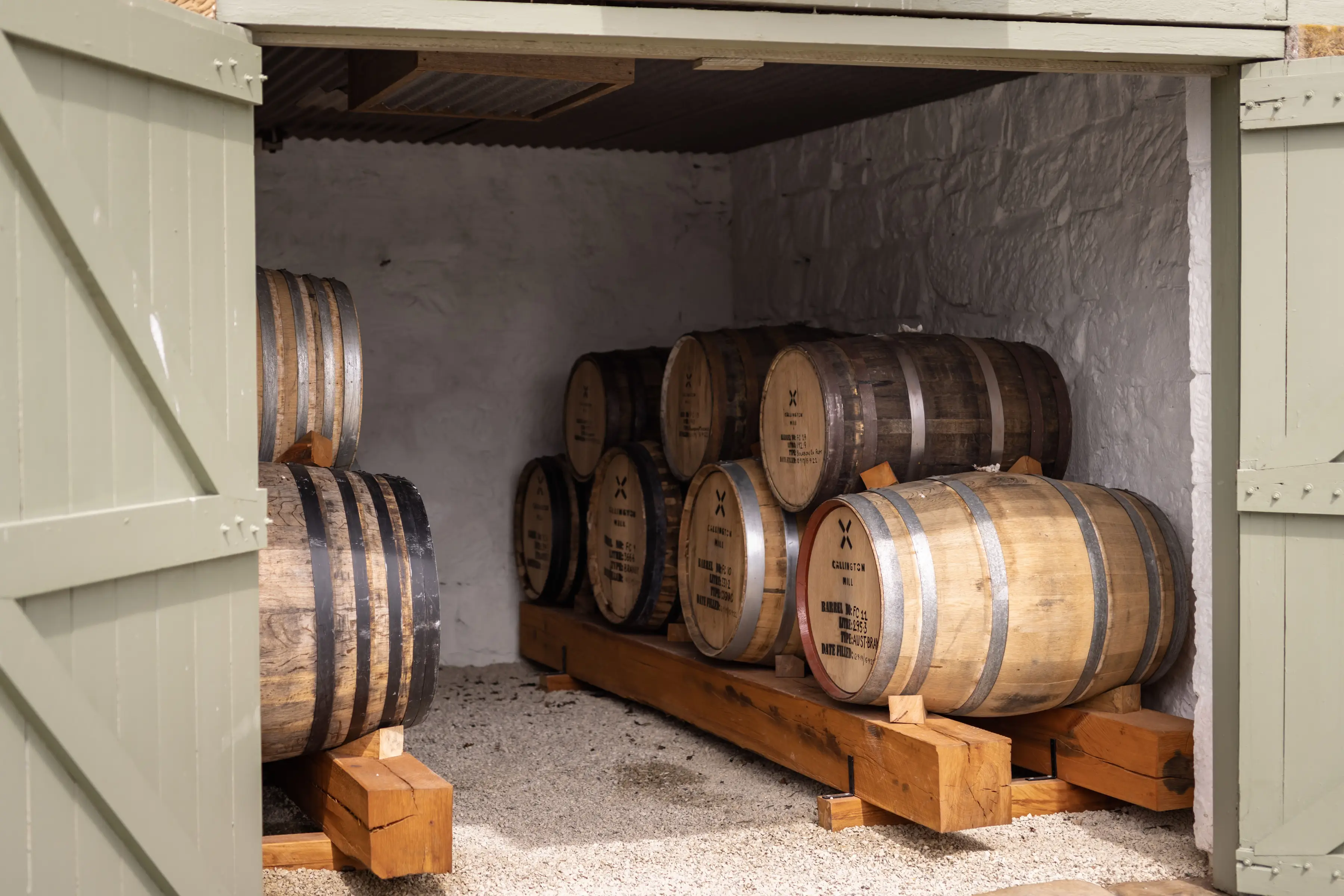 A small stone-walled cellar with rows of wooden aging barrels stacked on timber racks, viewed through open green doors. The barrels are marked with distillery labels and sit on a gravel floor beneath a corrugated metal ceiling.