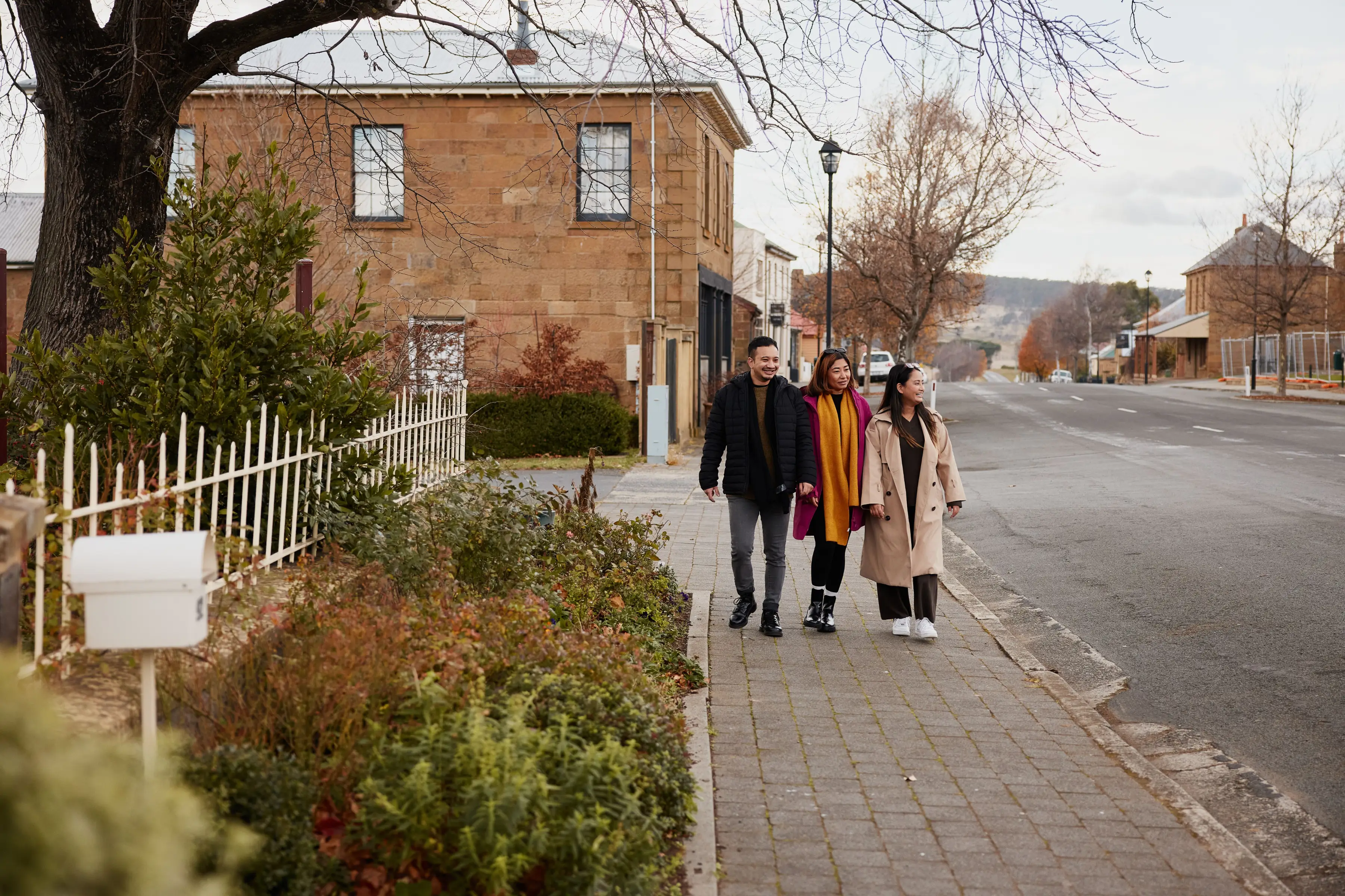 Three visitors stroll along a quiet heritage streetscape lined with historic sandstone buildings in Oatlands, Tasmania. Trees, gardens and colonial architecture showcase the village&rsquo;s rich history and charm.