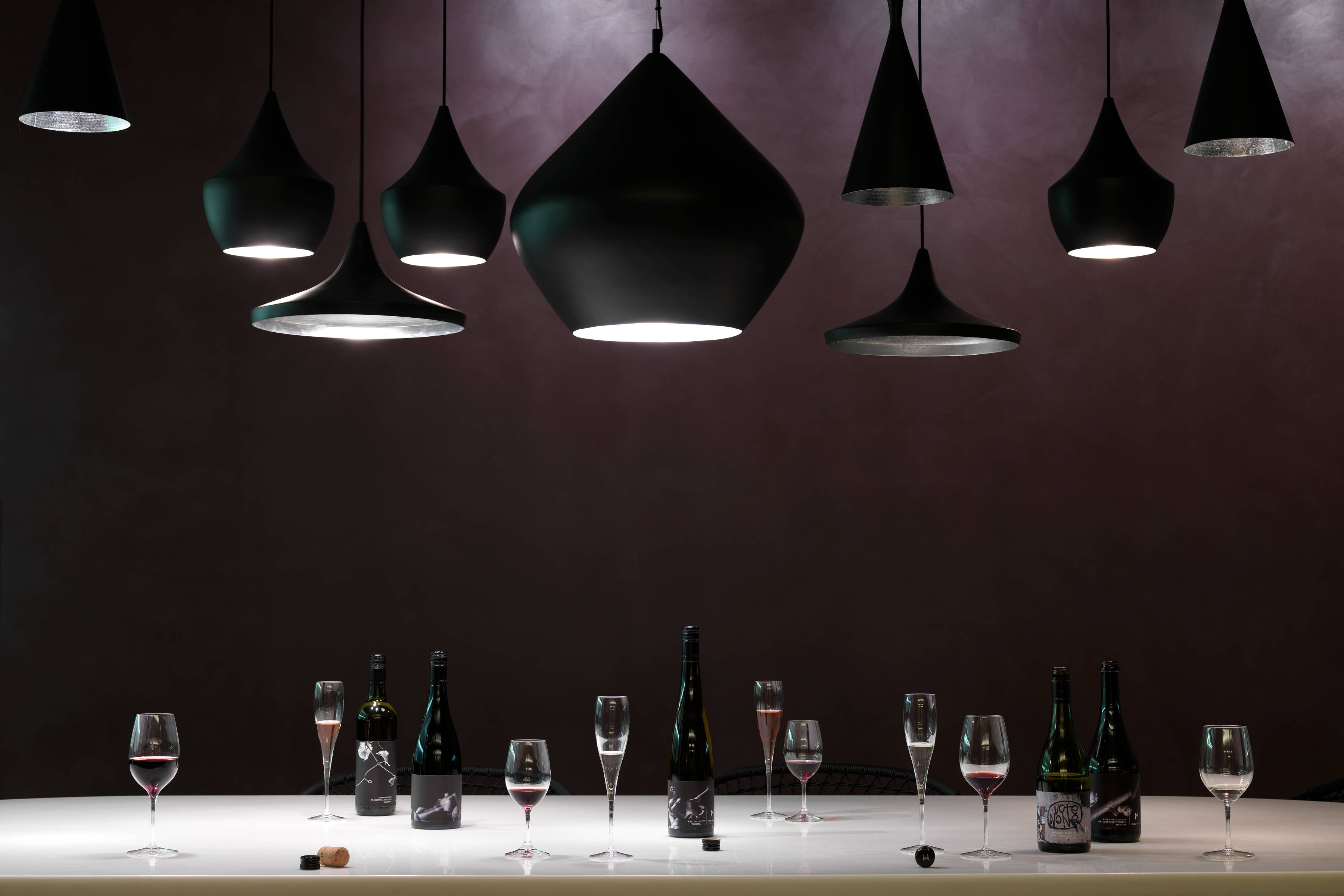 Moorilla’s wine tasting room featuring dark painted walls and dark pendant lights above the wine tasting table, on which five wine bottles and nine wine glasses sit.
