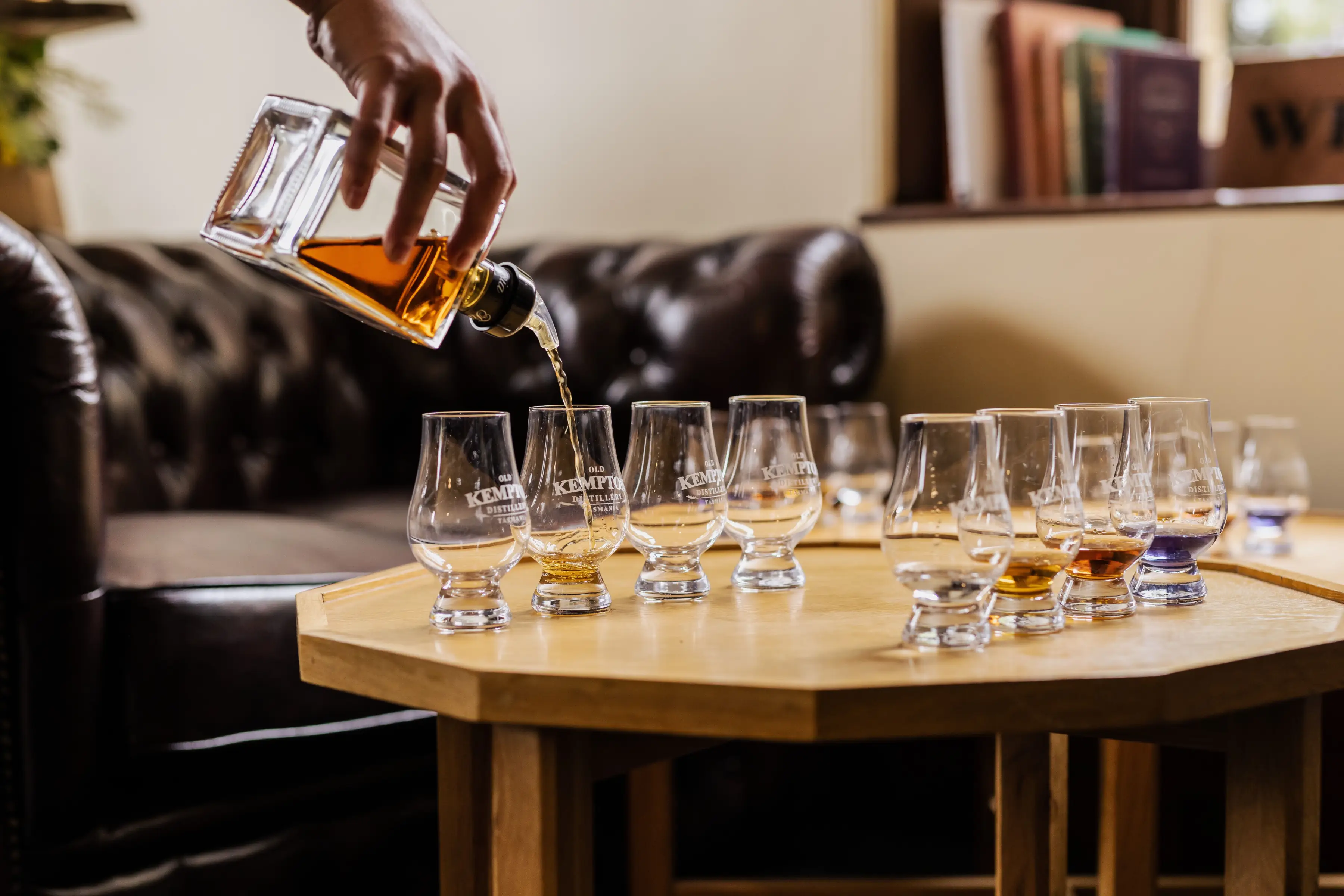 A hand pours golden-amber single malt whisky from a square glass bottle into a series of tasting glasses. The glasses sit on a light-coloured wooden table, with a leather Chesterfield couch visible in the soft-focus background.