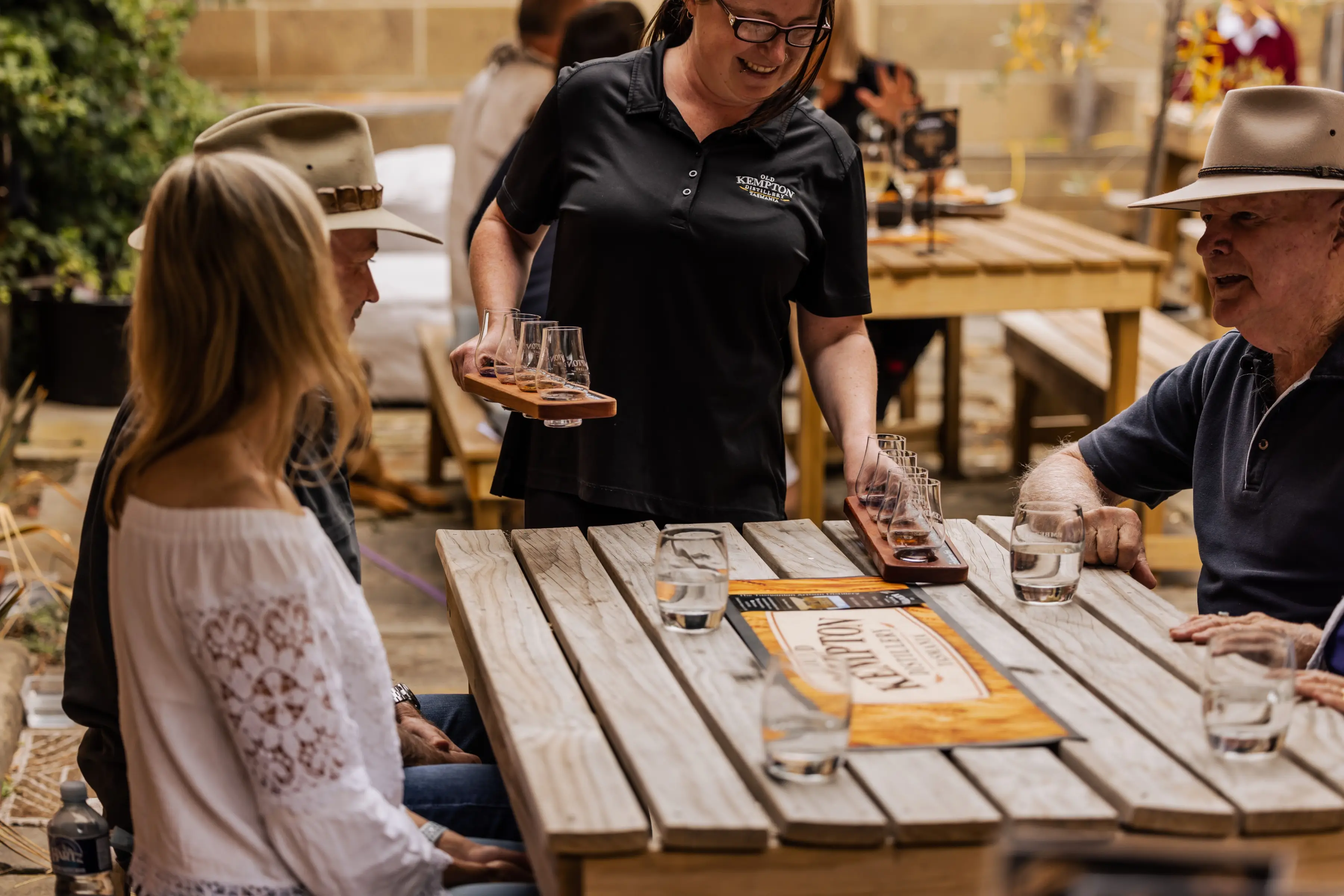 Guests enjoy a guided whisky tasting at an outdoor courtyard setting while a distillery host serves tasting flights. Wooden tables, tasting glasses and relaxed conversation create a welcoming and authentic Tasmanian experience.