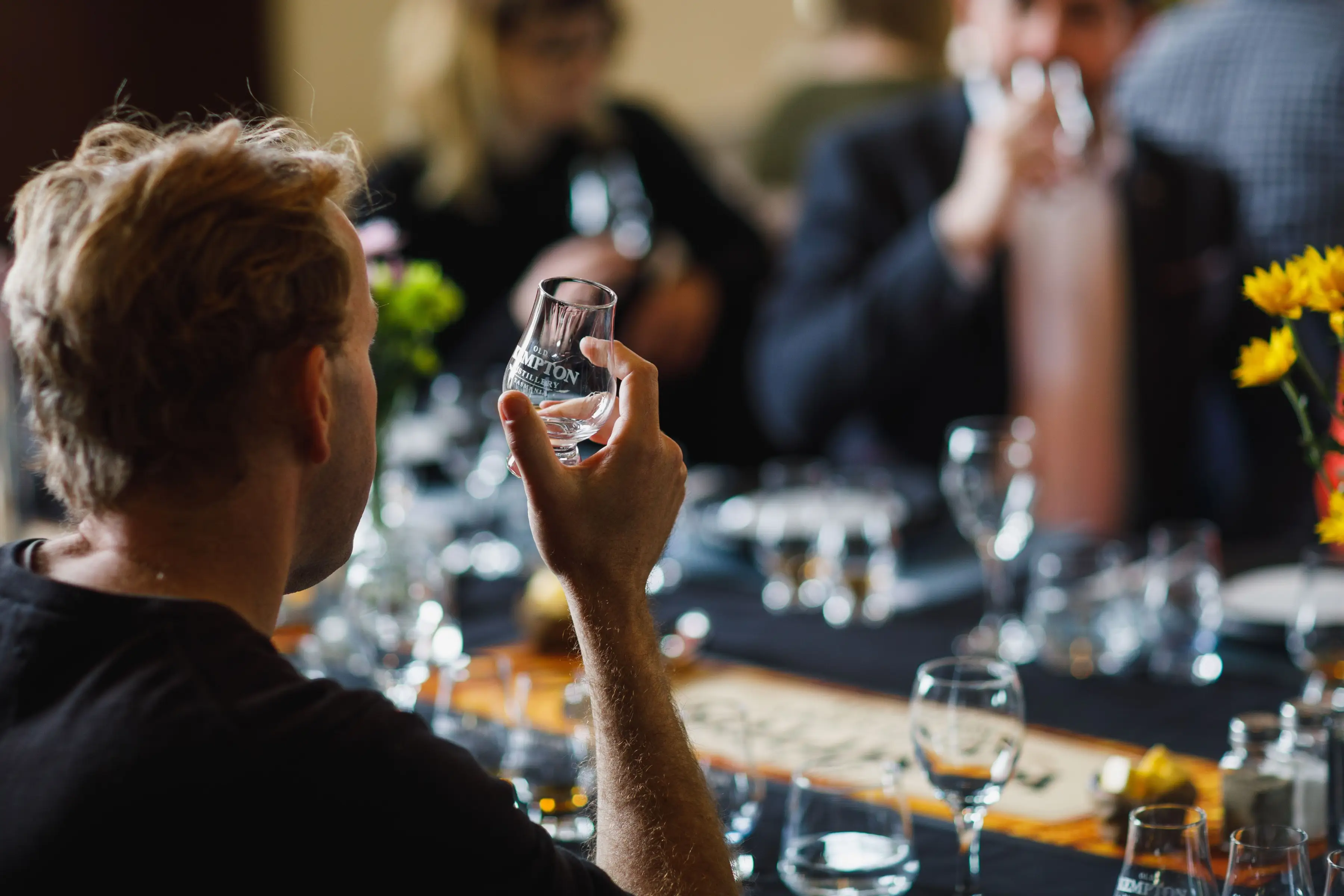 A guest holds a whisky tasting glass while seated at a tasting table during a guided distillery experience. Several tasting glasses are arranged on the table, with other participants softly blurred in the background, creating a warm and social atmosphere.