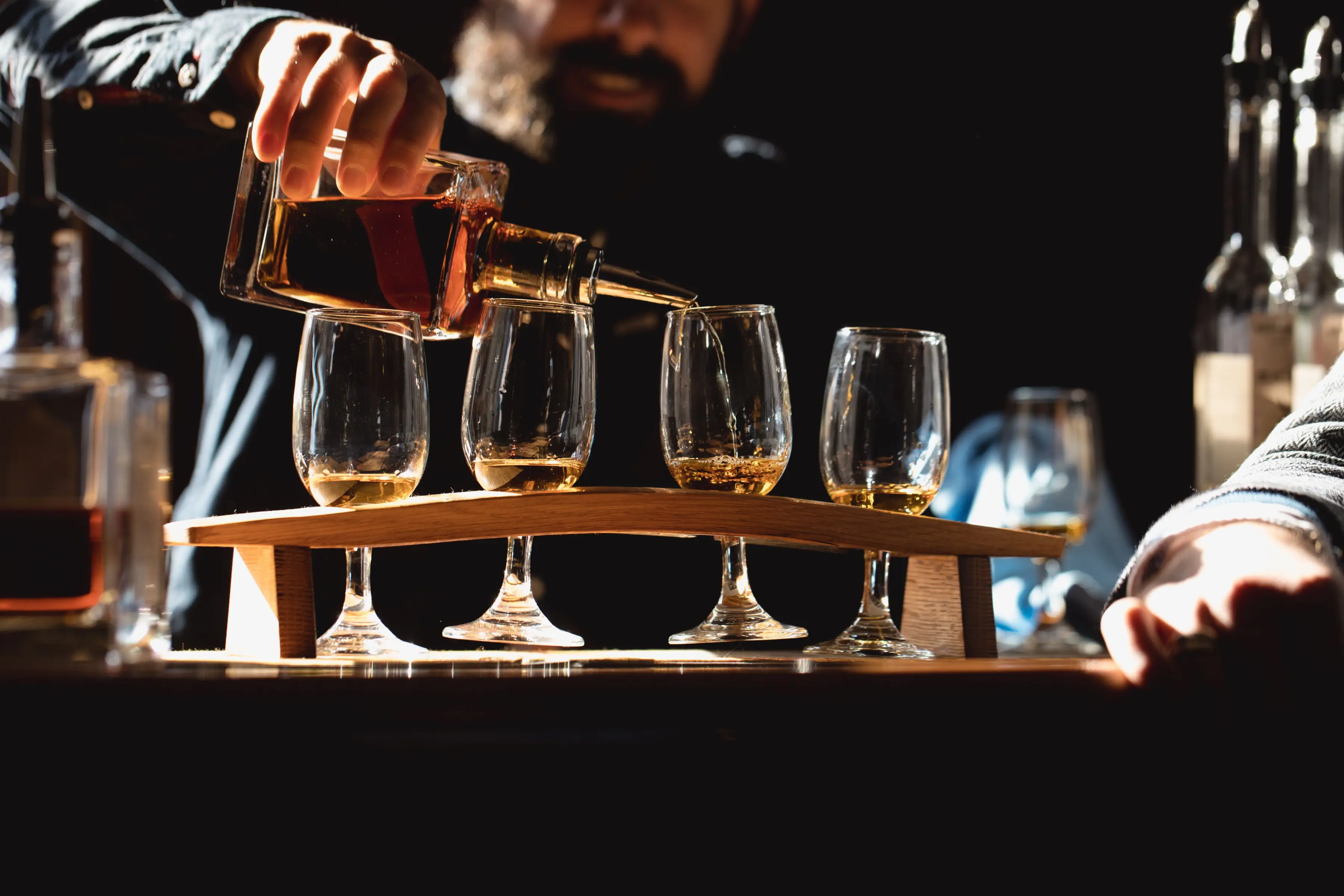 A distillery staff member pours spirit from a glass bottle into a flight of tasting glasses arranged on a wooden tasting paddle. Warm lighting highlights the golden liquid and creates an inviting tasting room atmosphere.