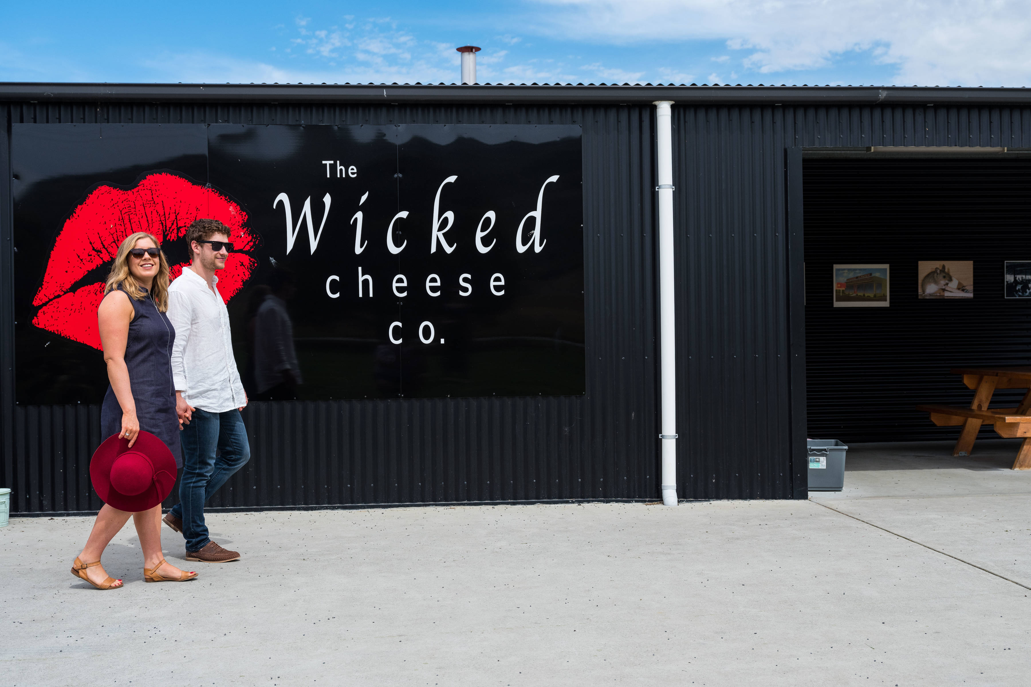 Wicked Cheese front entrance. The Wicked Cheese building is painted black with large black signage featuring the Wicked Cheese logo, consisting of red lips and white lettering. A man and a woman are walking in front of the sign.