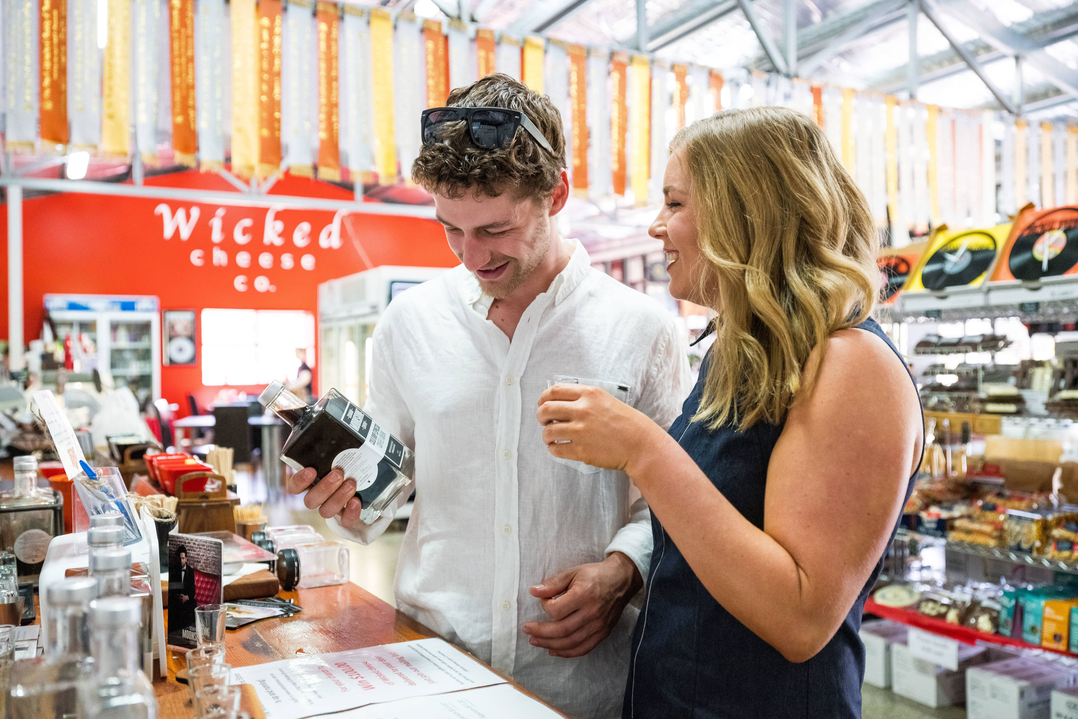 A man and a woman inside the Wicked Cheese shop. The man is reading the label on a bottle of cold drop espresso coffee liqueur and the woman is tasting a sample.